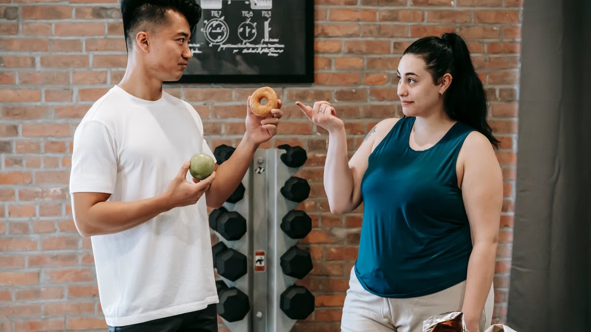 Side view of Asian personal instructor with apple and doughnut offering healthy food to plus size woman while standing in gym